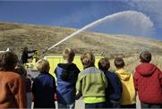 A group of adults watching firemen spray a water hose from a fire truck.